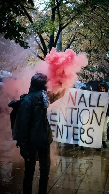 Demonstranten met rode rookfakkels lopen achter een spandoek tegen detentiecentra